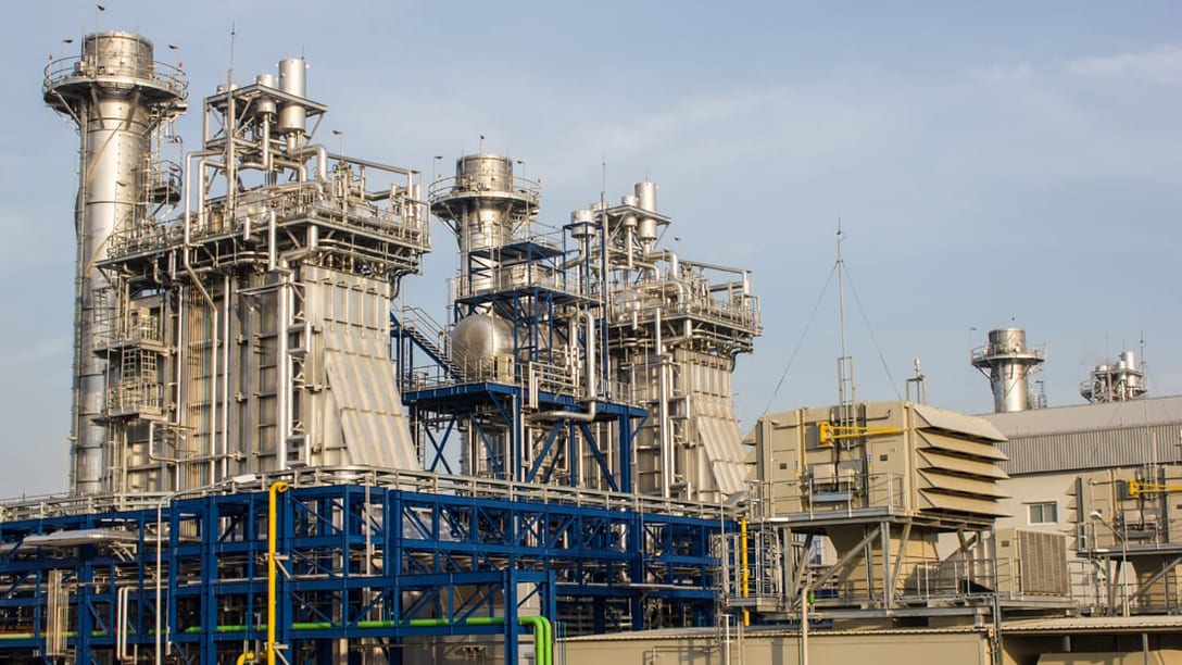 Blue pipe racks and skid-mounted equipment in front of a combined-cycle plant's heat-recovery units and stacks